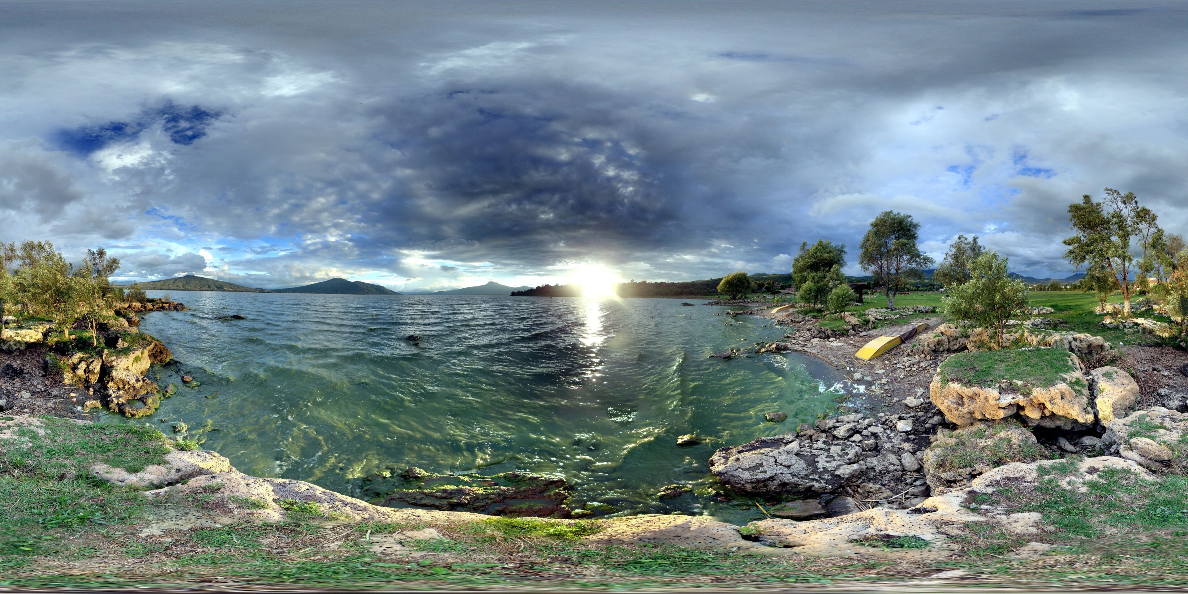 A panoramic view of a lake with clear water, surrounded by rocky shores and trees, under a cloudy sky.