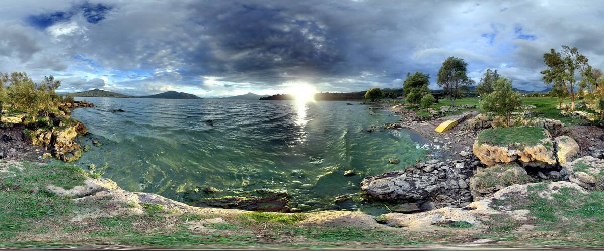 A panoramic view of a lake with clear water, surrounded by rocky shores and trees, under a cloudy sky.