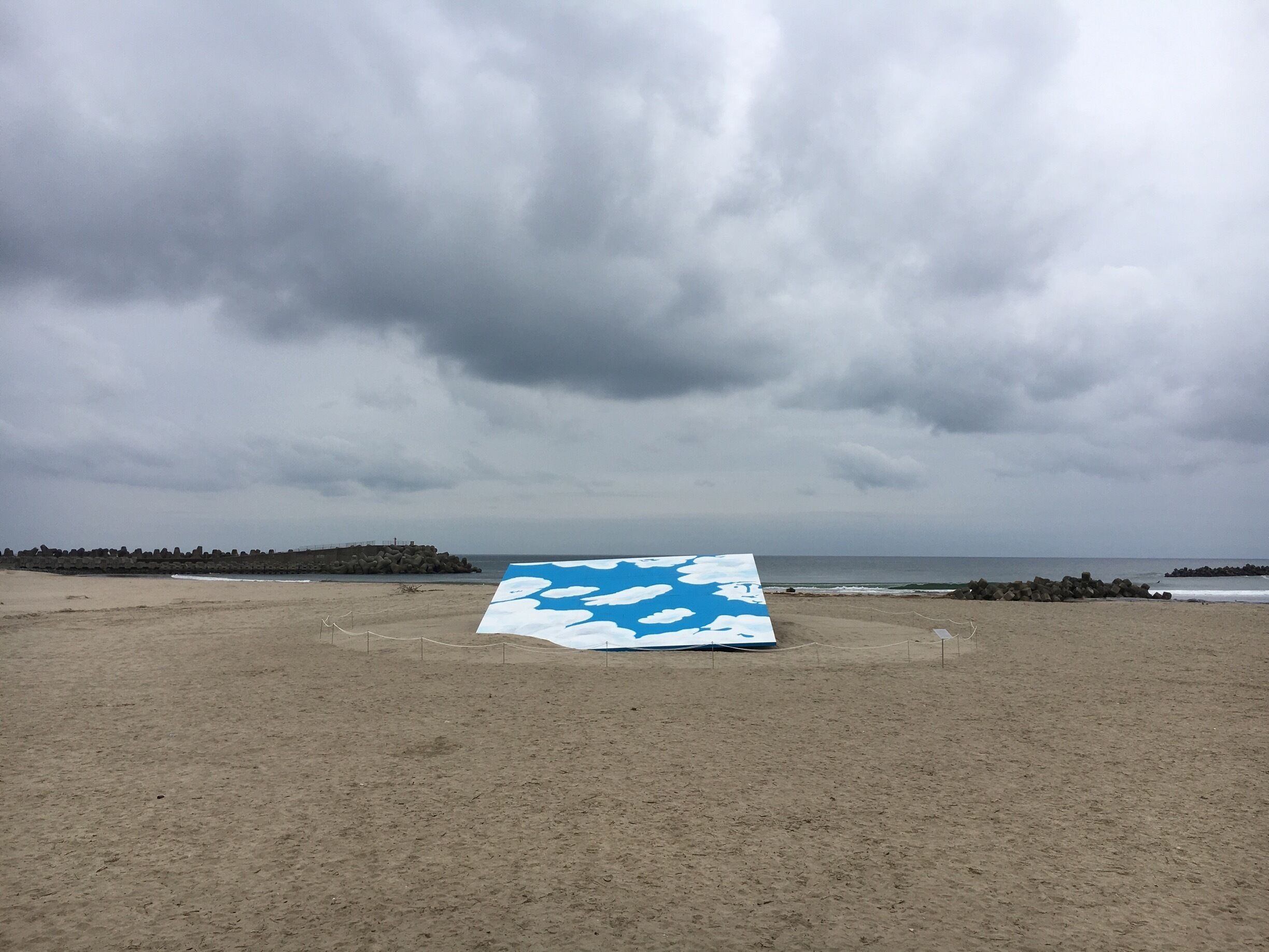 A beach with a large blue and white canvas on the sand, a rocky breakwater, and the ocean in the background.