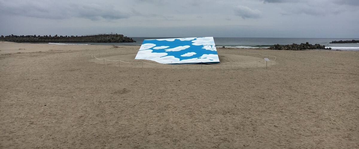 A beach with a large blue and white canvas on the sand, a rocky breakwater, and the ocean in the background.