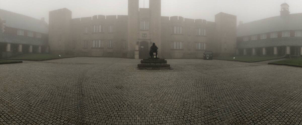 A foggy courtyard with a statue in the center, surrounded by buildings with multiple windows and a tower.