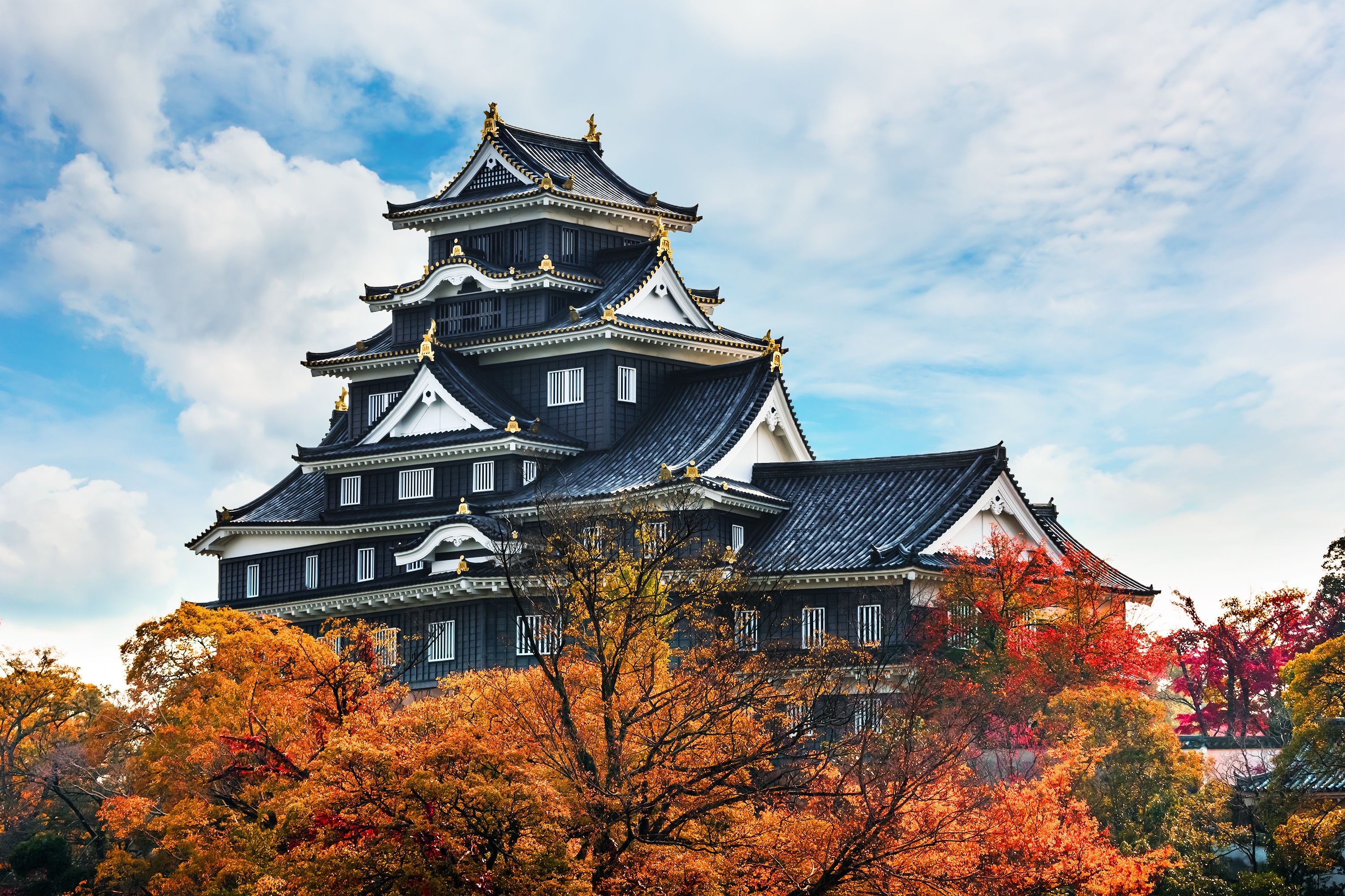 A traditional Japanese castle with multiple tiers and golden decorations, surrounded by autumn-colored trees.