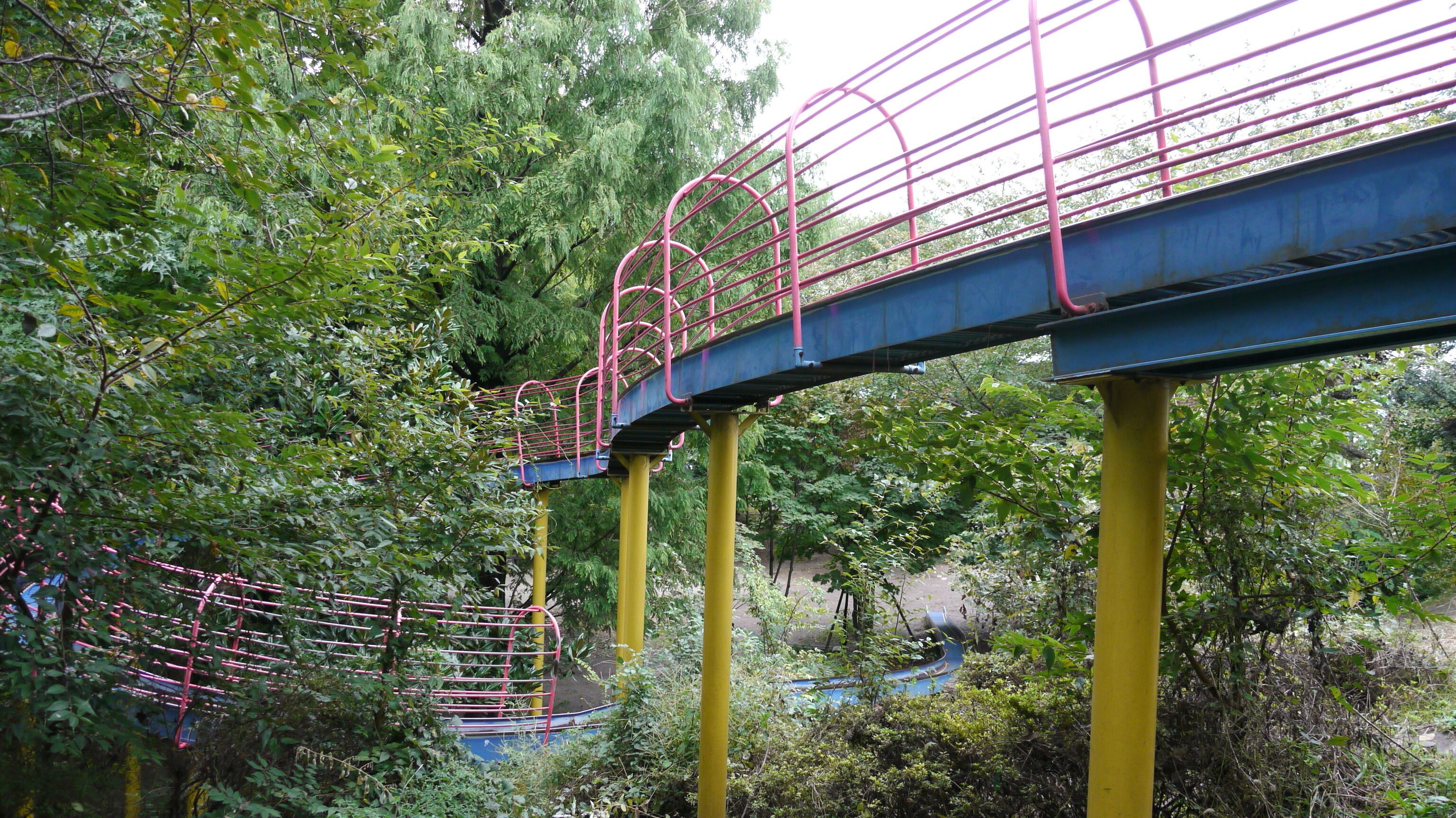 A metal bridge with pink railings, supported by yellow pillars, surrounded by dense green foliage.