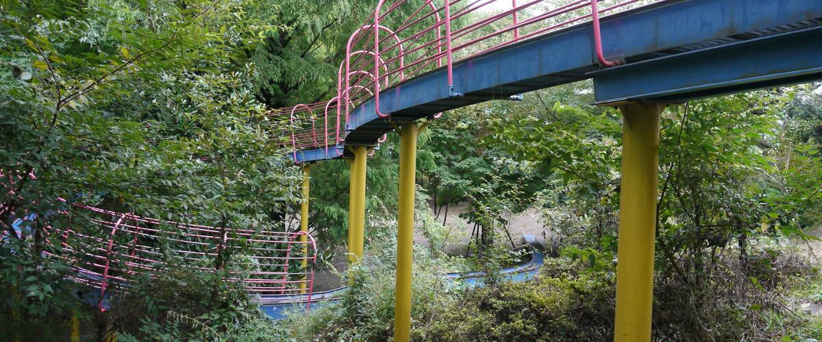 A metal bridge with pink railings, supported by yellow pillars, surrounded by dense green foliage.