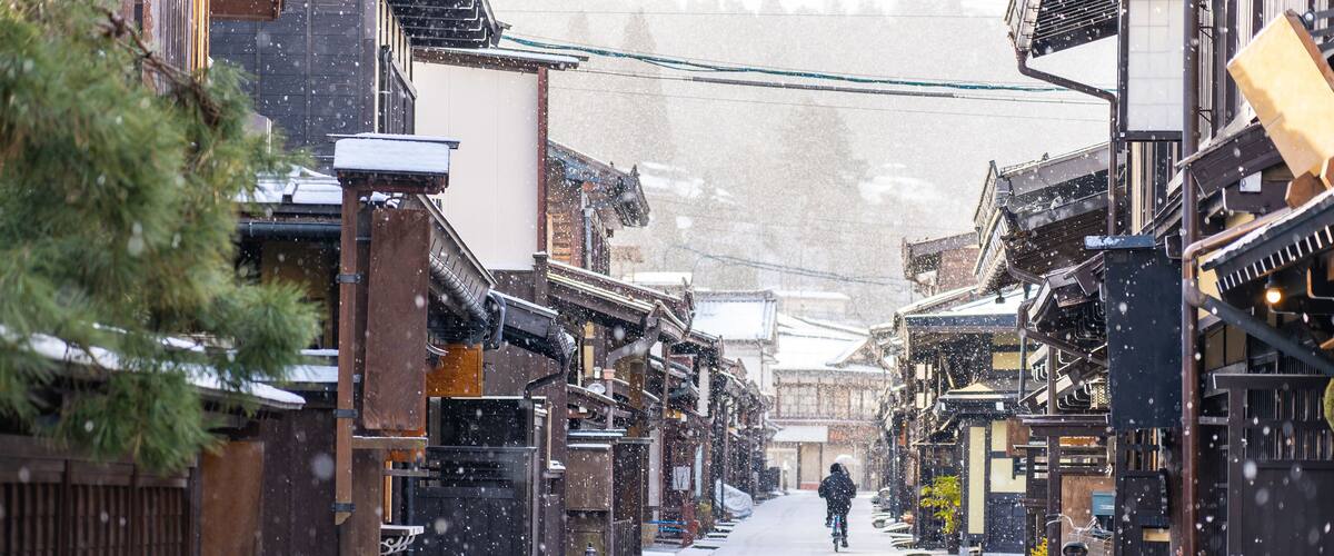 A snow-covered street in a traditional town with wooden buildings and a person riding a bicycle.
