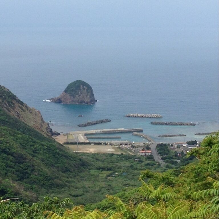A coastal landscape with a prominent green hill, a small island with a rocky outcrop, and a series of breakwaters extending into the sea.