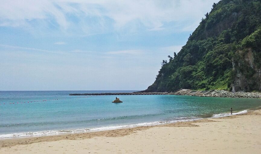 A beach with clear turquoise water, a rocky outcrop, and a person walking along the shore.