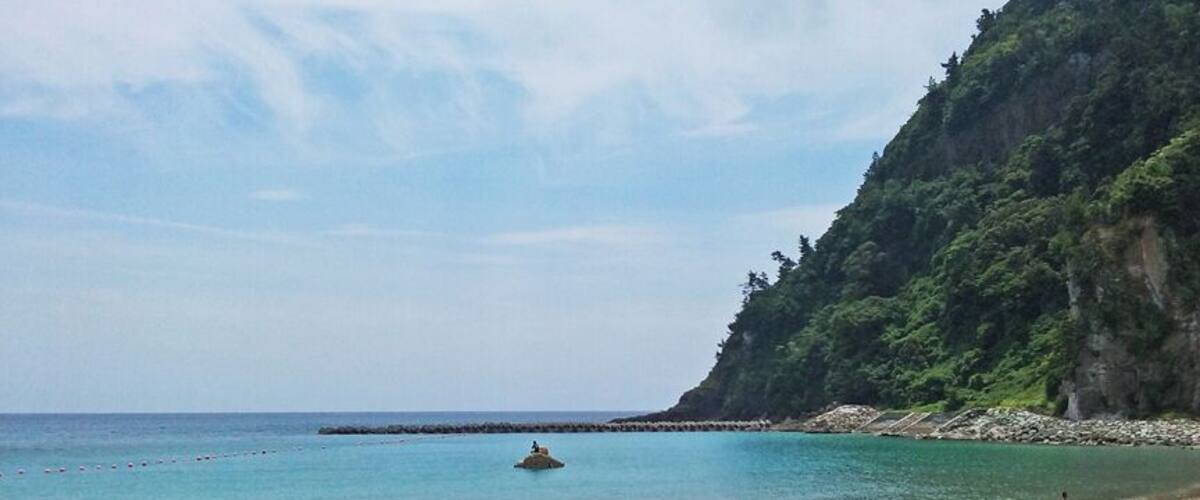 A beach with clear turquoise water, a rocky outcrop, and a person walking along the shore.