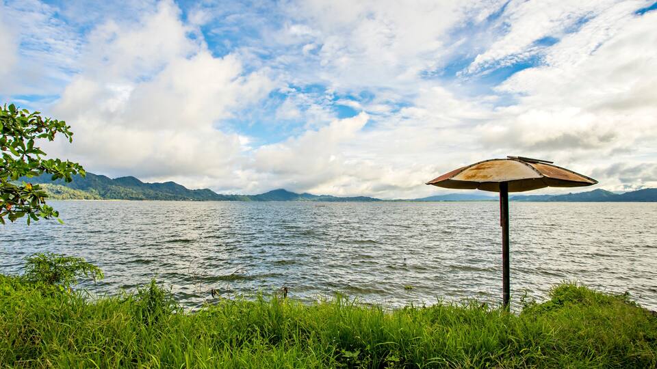 A lake with a rusty umbrella on the shore, surrounded by greenery and mountains in the background.