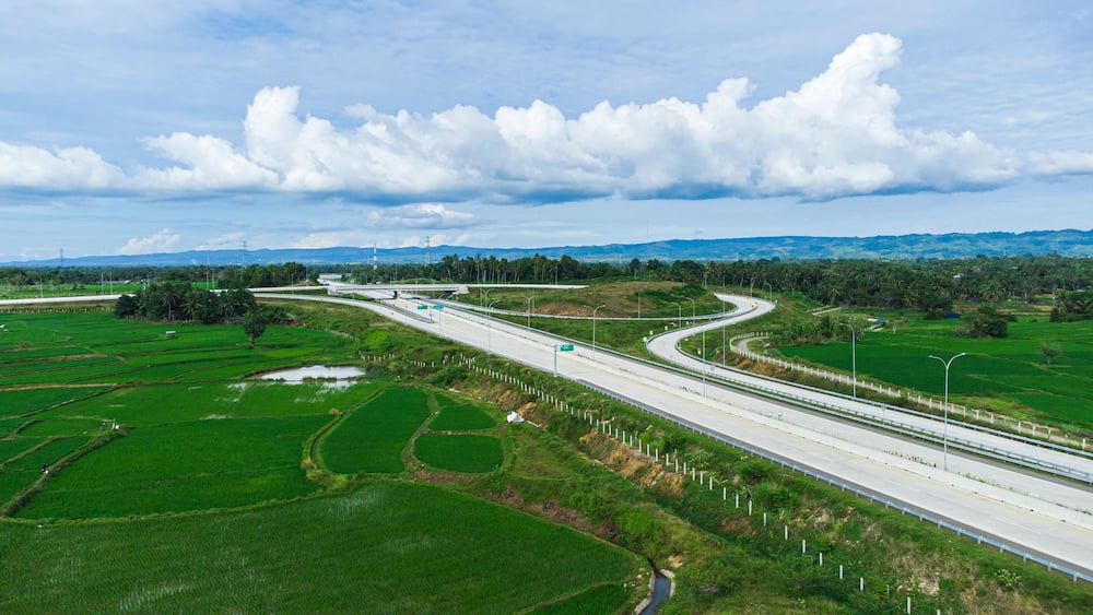 A highway curving through green fields with distant mountains and a cloudy sky.