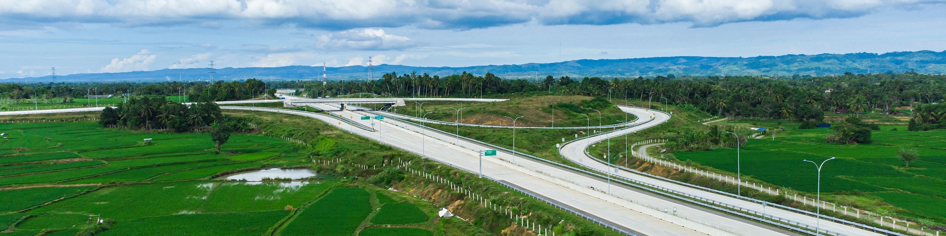 A highway curving through green fields with distant mountains and a cloudy sky.