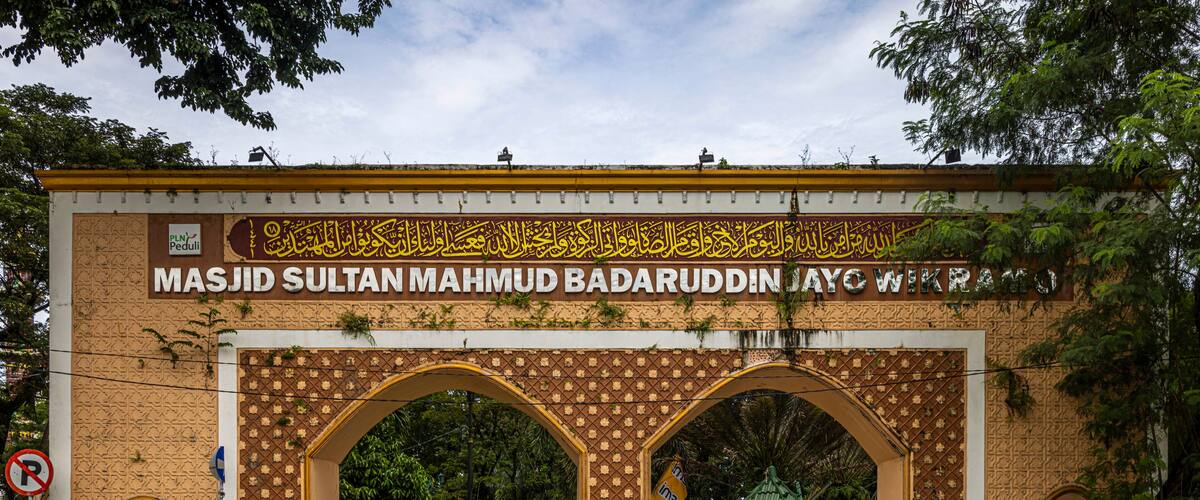 A large archway with intricate patterns and Arabic calligraphy, leading to a building with a green roof.