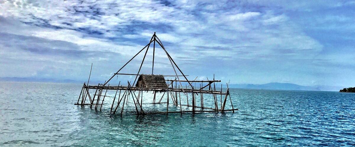 A traditional wooden raft floating on calm water with a thatched roof, surrounded by a clear sky and distant mountains.