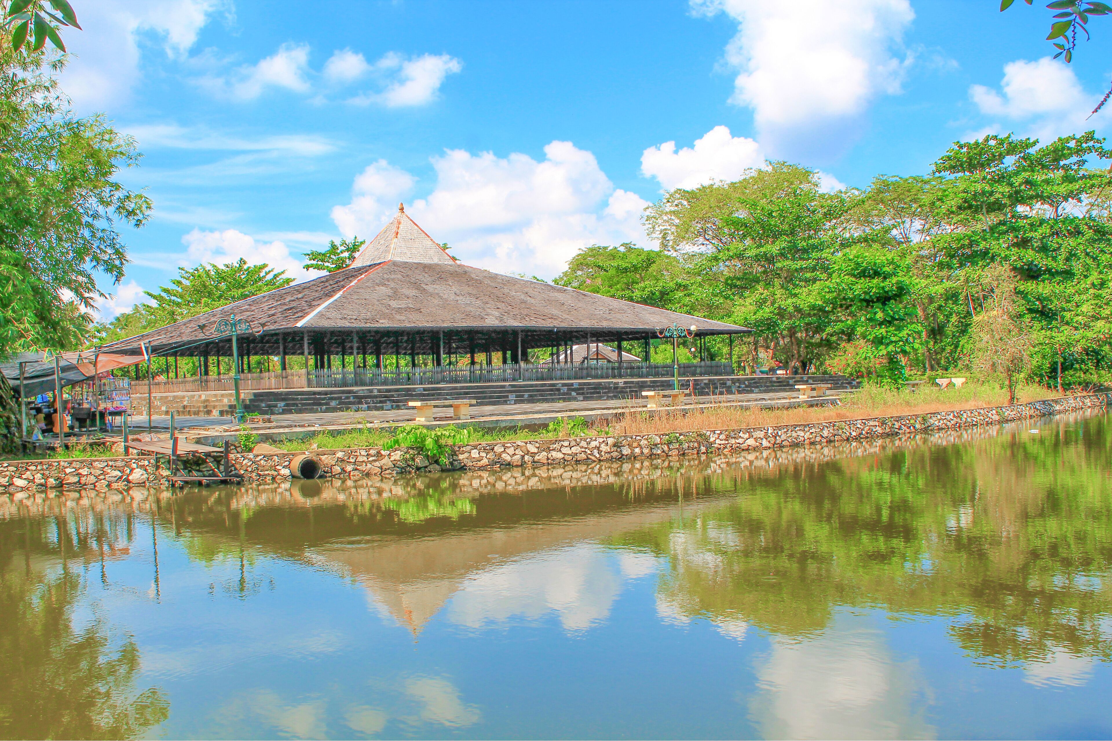 A pavilion with a thatched roof by a calm pond, surrounded by lush greenery and a clear sky.