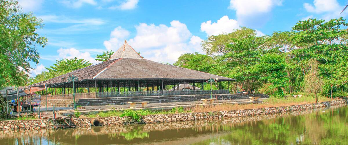 A pavilion with a thatched roof by a calm pond, surrounded by lush greenery and a clear sky.