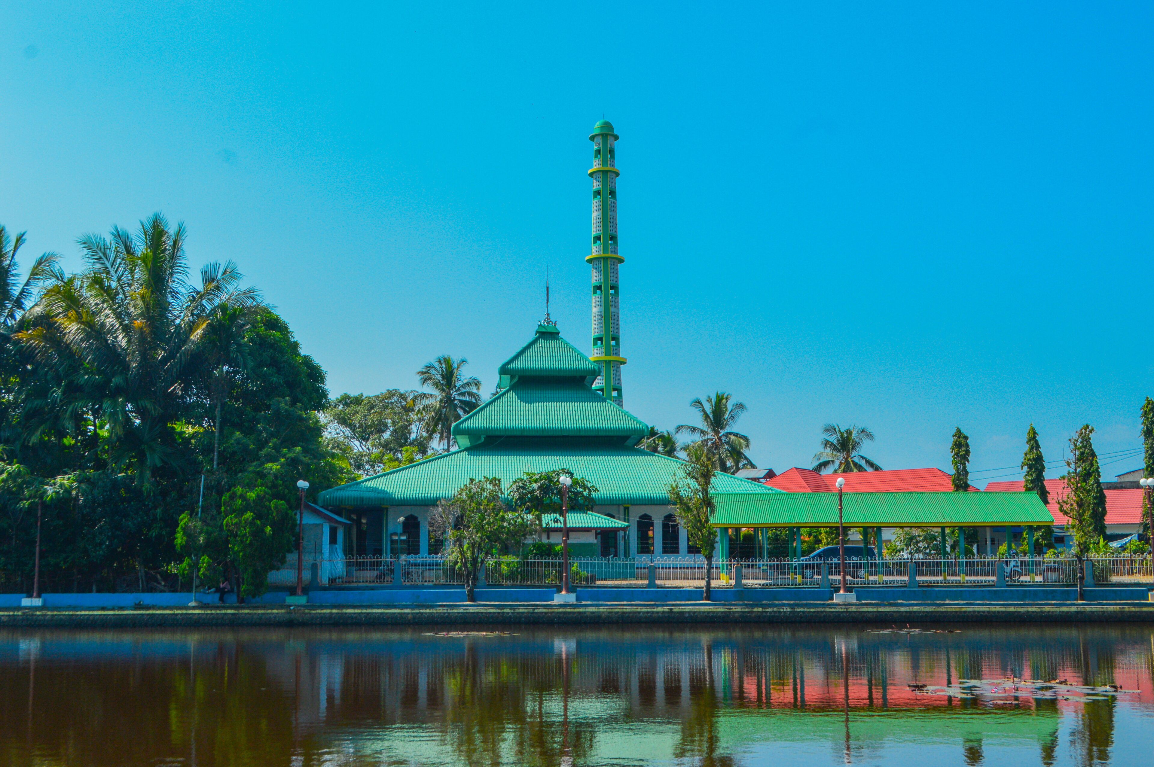 A mosque with a green dome and minaret by a calm lake, surrounded by palm trees and a red-roofed building.
