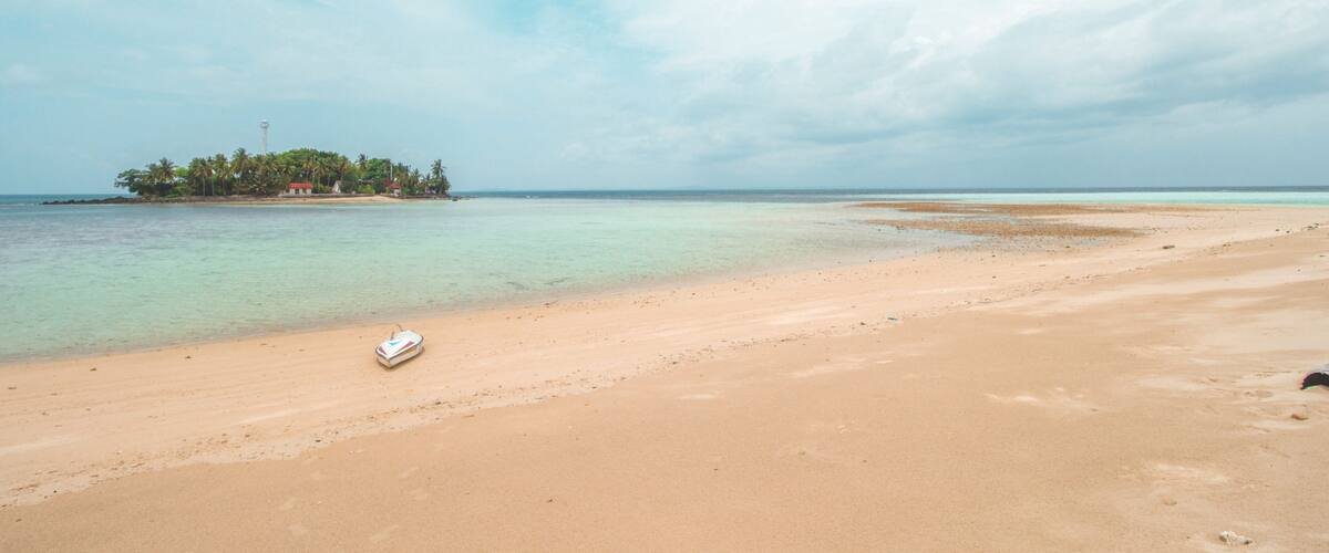 A beach with clear turquoise water, a small island with palm trees, and a boat on the sand.