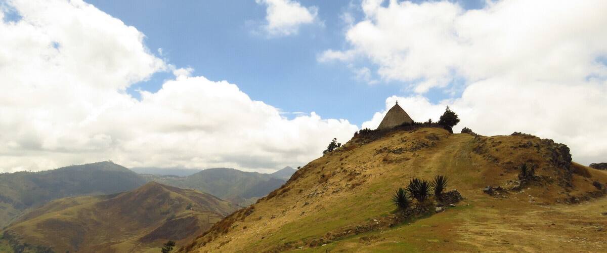 A grassy hill with a thatched-roof structure on top, surrounded by rolling hills and a partly cloudy sky.