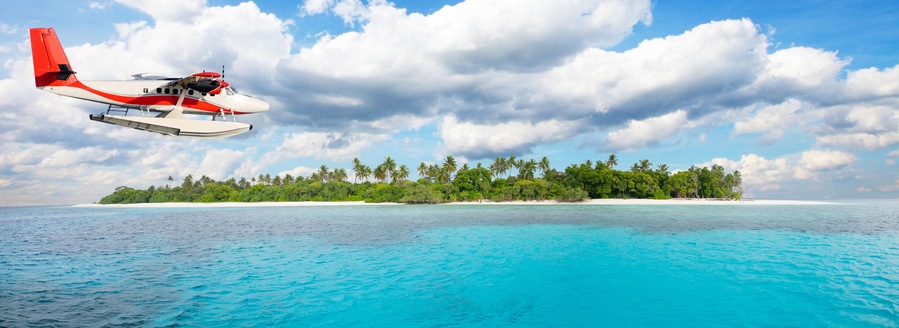 A seaplane flying over turquoise waters with a tropical island in the background.