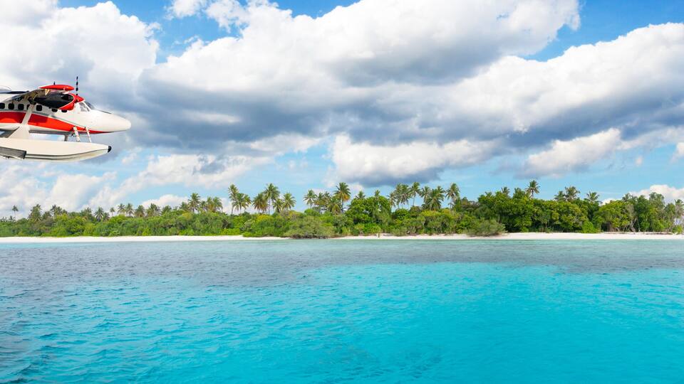 A seaplane flying over turquoise waters with a tropical island in the background.
