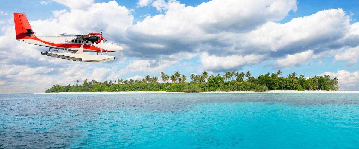 A seaplane flying over turquoise waters with a tropical island in the background.