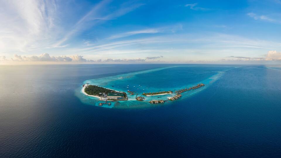 An aerial view of a tropical island with clear turquoise waters, a central building complex, and a small peninsula with a dock.