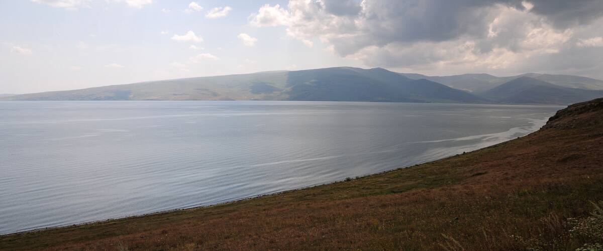 A calm lake with a grassy shoreline and distant mountains under a cloudy sky.