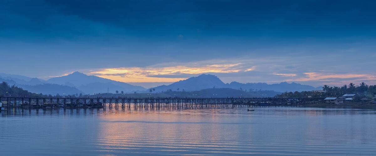 sunrise at Sangkhlaburi, river view morning on the river of old wooden bridge (Mon Bridge) with reflection on the water, mountain and colorful of yellow sun light in the sky, Kanchanaburi, Thailand.