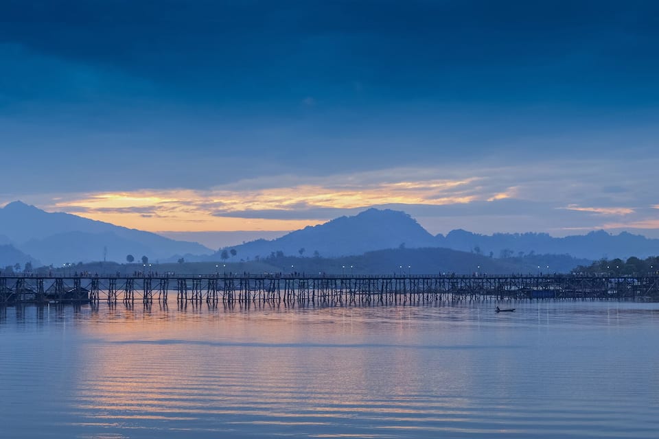 Um lago calmo com uma ponte de madeira, montanhas ao fundo e um pôr do sol.
