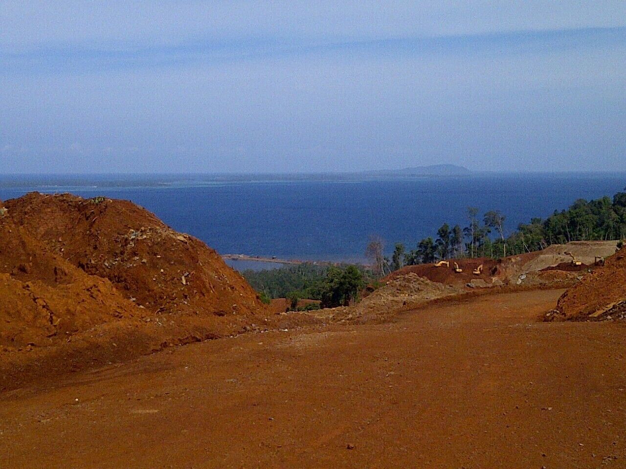 A barren landscape with reddish soil, construction equipment, and a distant view of the ocean and a mountain range.