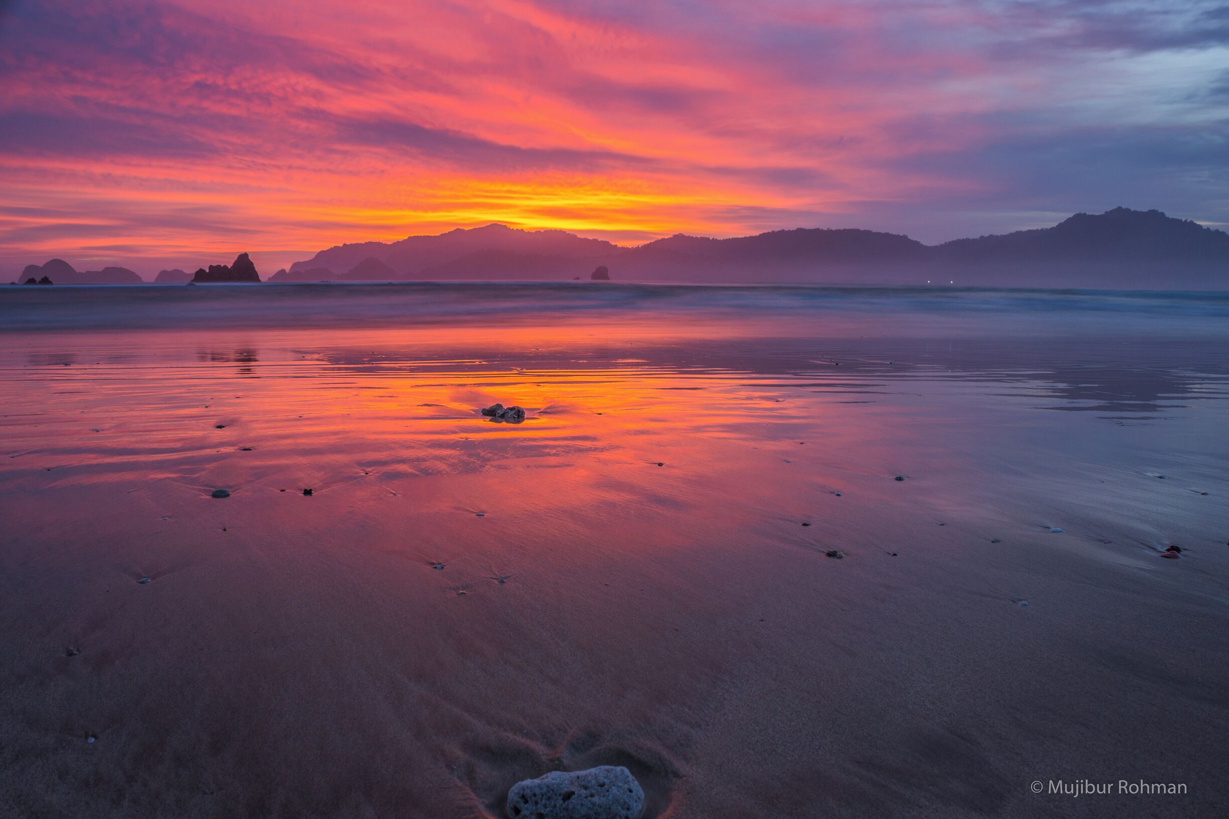 A beach at sunset with a colorful sky, calm water, and distant mountains.