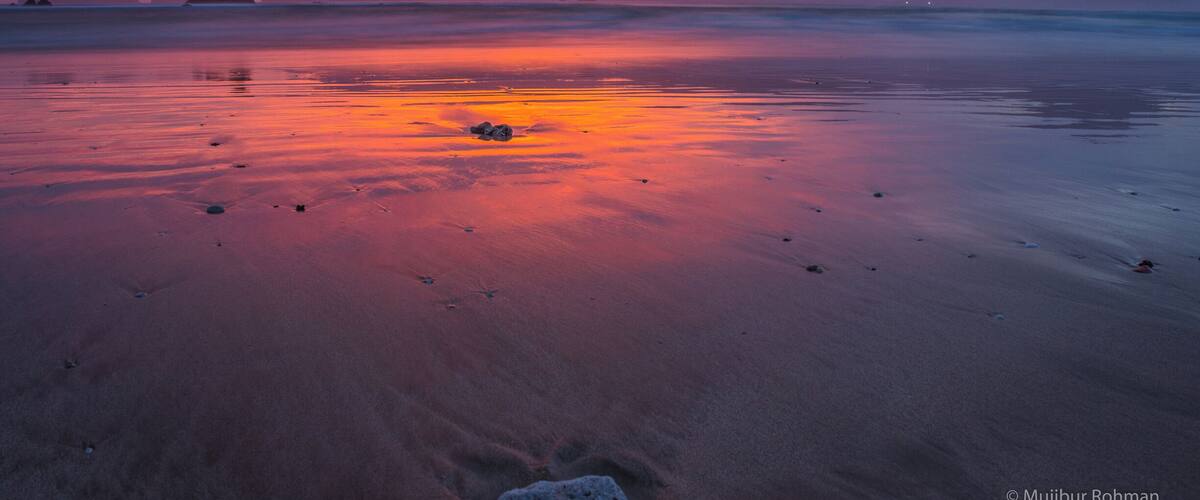 A beach at sunset with a colorful sky, calm water, and distant mountains.