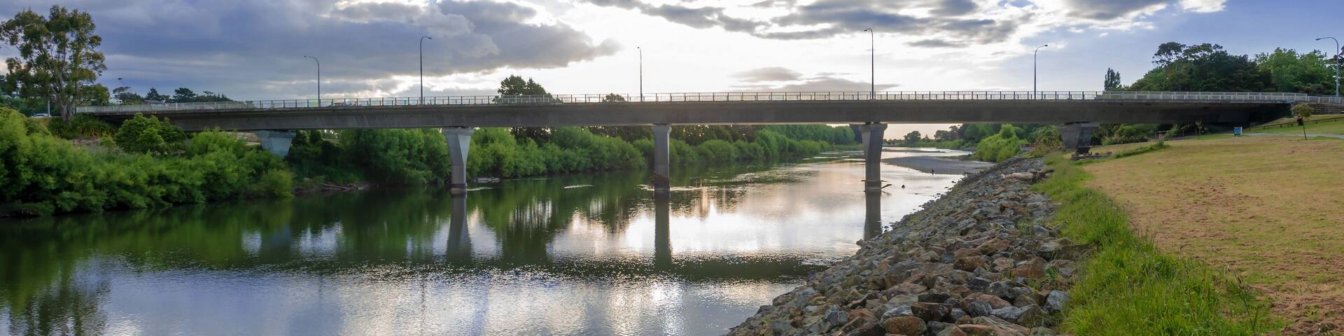 A river with a rocky embankment, a bridge, and a cloudy sky.