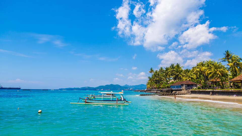 A tropical beach with clear turquoise water, a few boats, and palm trees.