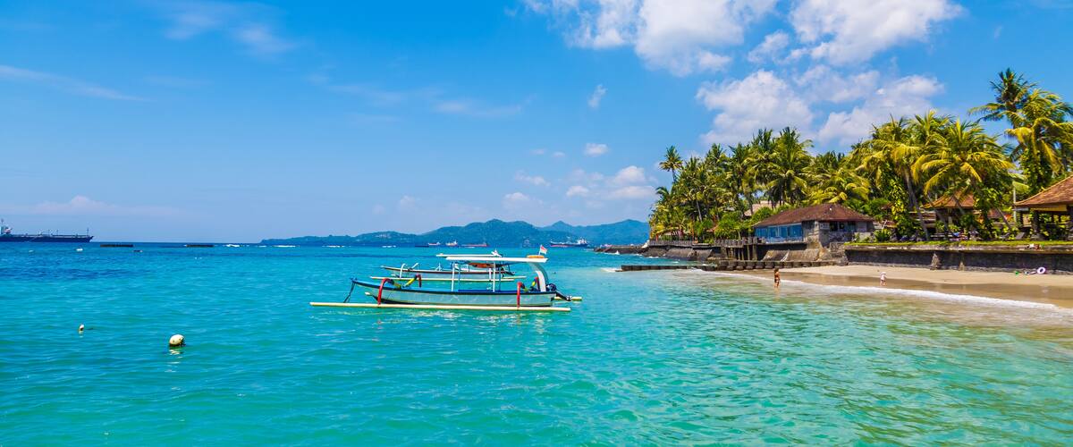 A tropical beach with clear turquoise water, a few boats, and palm trees.