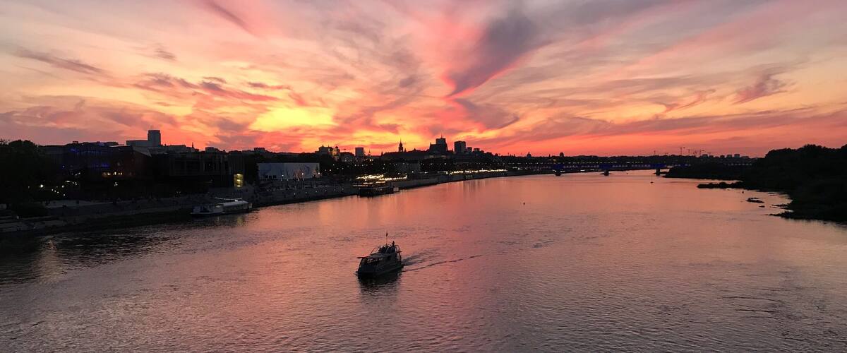A sunset over a river with a boat in the foreground and a city skyline in the background.