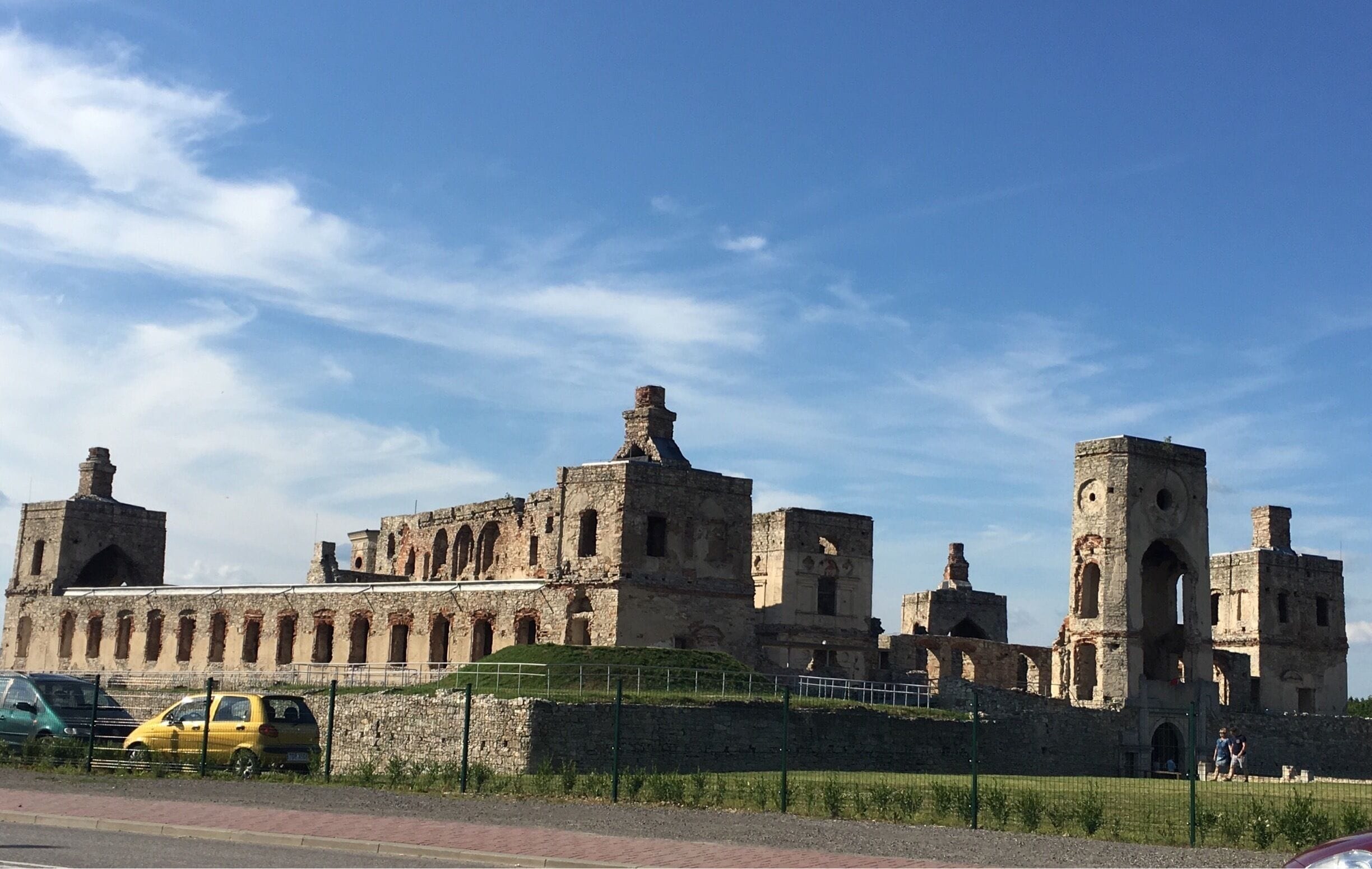 An ancient stone fortress with multiple towers and arched windows, surrounded by a green fence and a clear blue sky.