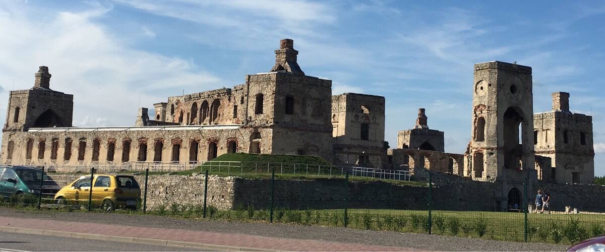 An ancient stone fortress with multiple towers and arched windows, surrounded by a green fence and a clear blue sky.