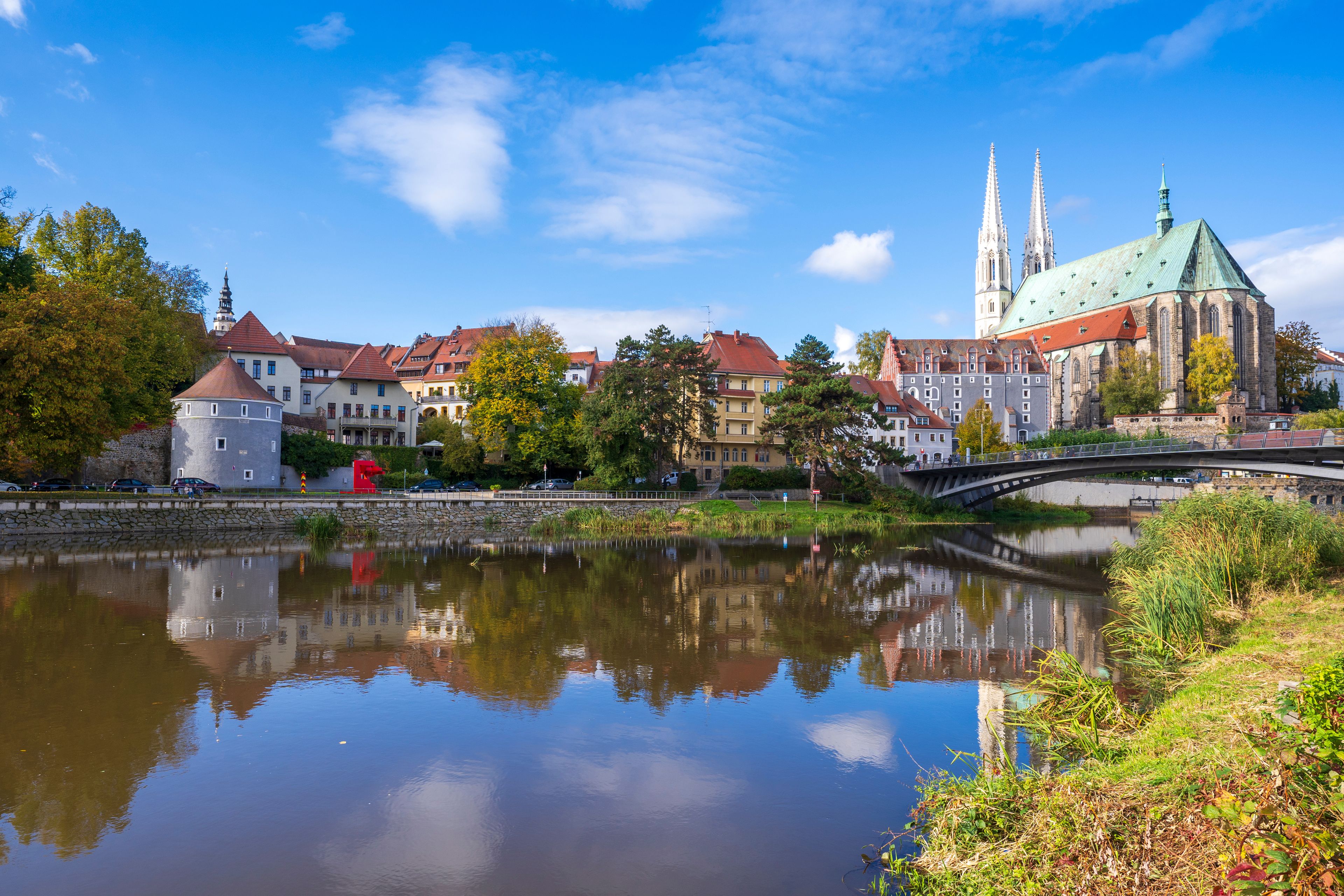 A river reflecting buildings, a bridge, and a church with twin spires.