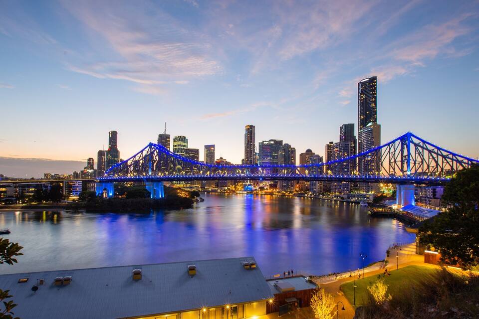 A city skyline with a prominent bridge illuminated in blue, reflecting on the water.