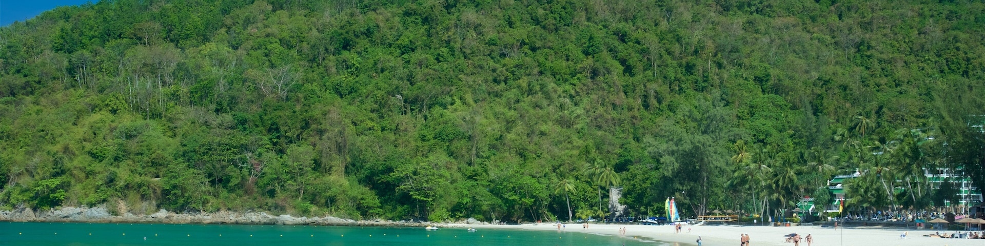 Ein Strand mit klarem türkisblauem Wasser, einem sandigen Ufer und einem bewaldeten Hügel im Hintergrund.