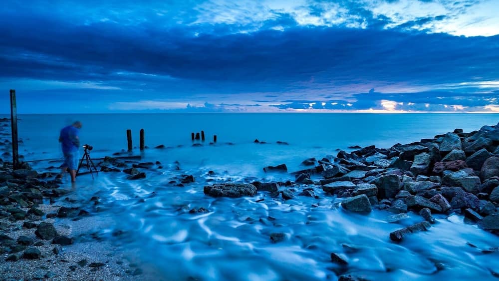 A rocky shoreline with a person standing near a tripod, a calm sea, and a sunset sky.