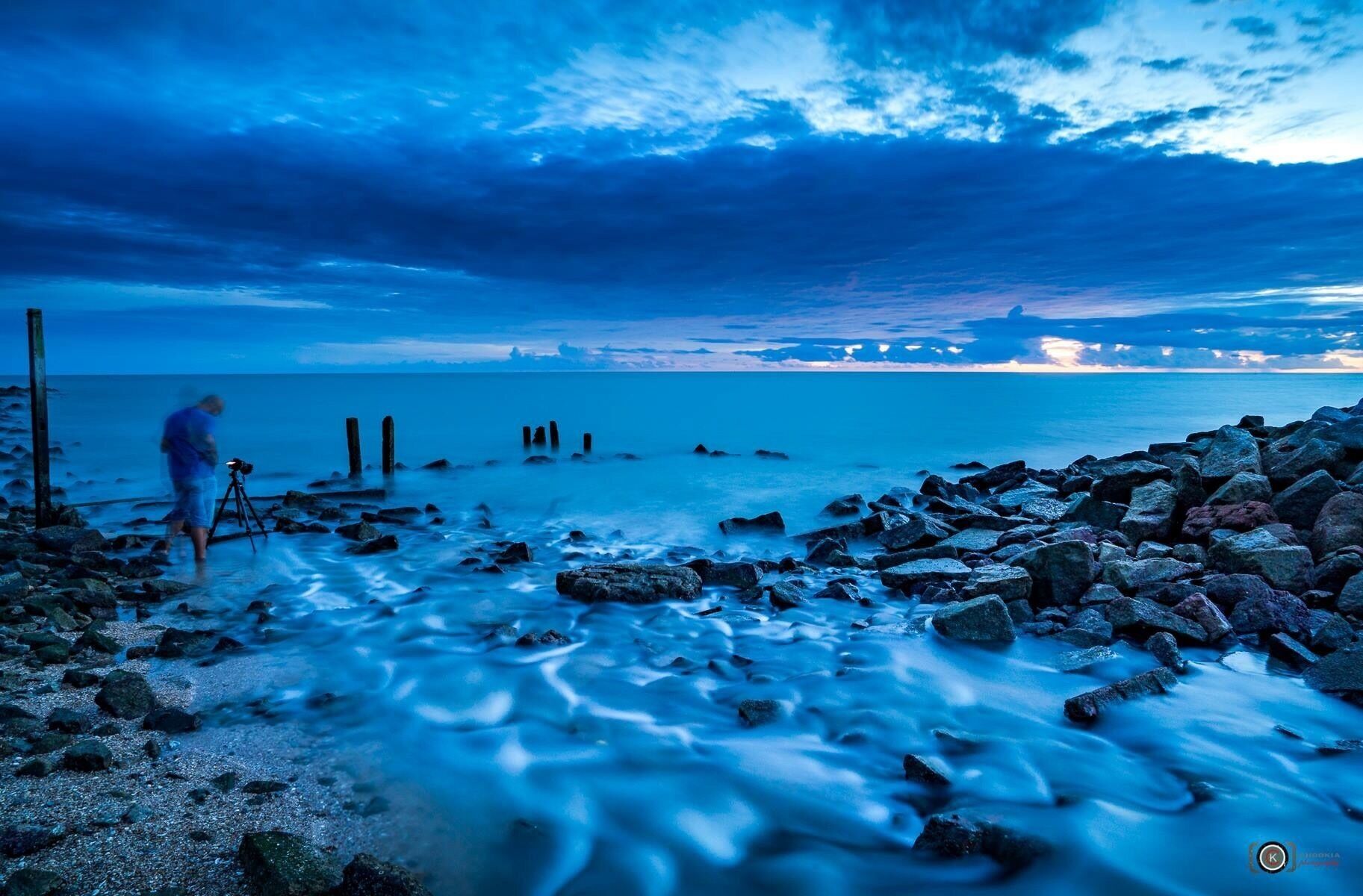 A rocky shoreline with a person standing near a tripod, a calm sea, and a sunset sky.