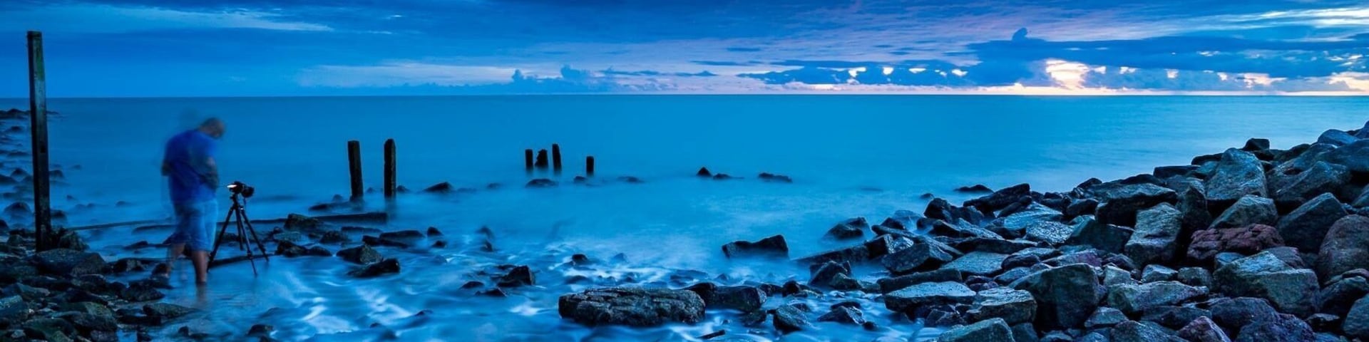 A rocky shoreline with a person standing near a tripod, a calm sea, and a sunset sky.