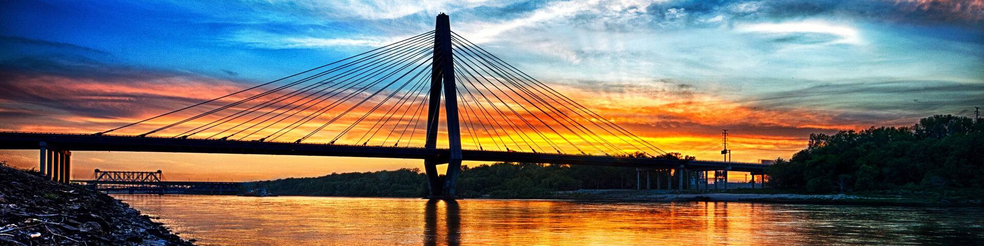 A suspension bridge at sunset with a rocky shoreline.