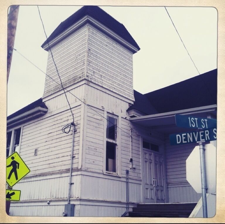 Um edifício branco com chaminé, sinal de faixa de pedestres e placas de rua indicando 1st St e Denver St.