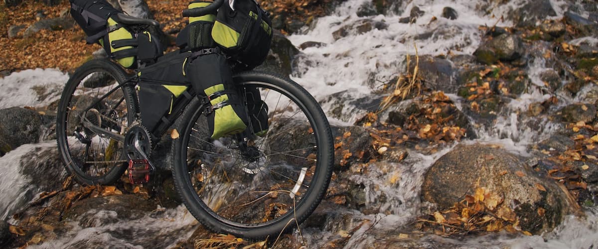 Una bicicleta de muntanya amb un cistell davanter i una arrencada al darrere, aparcada en un camí pedregós amb un bosc de fons.
