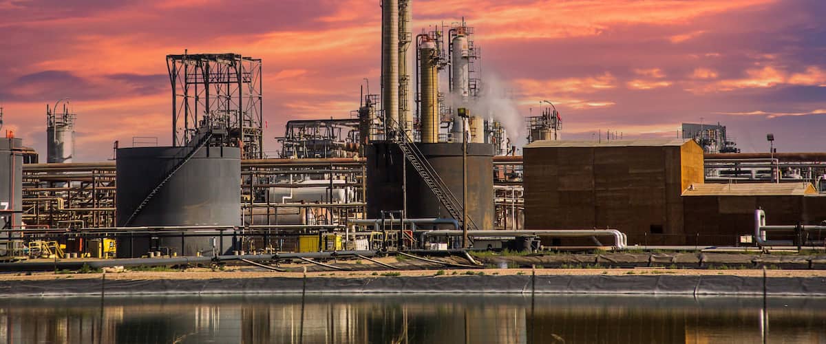 An industrial facility with tall chimneys and storage tanks, set against a sunset sky.