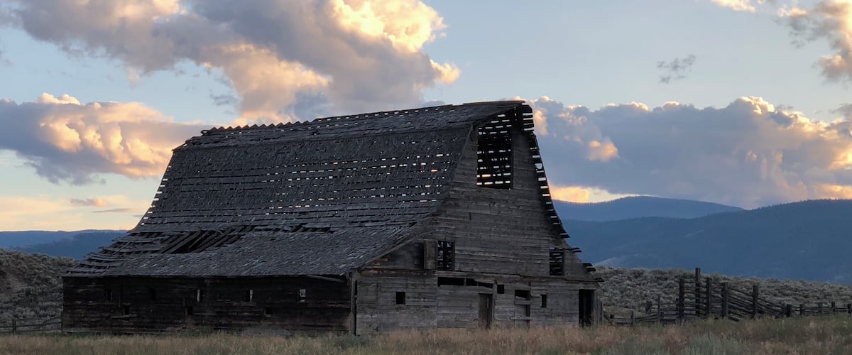 A dilapidated wooden barn with a steep roof, surrounded by a dry grassy field and mountains in the background.