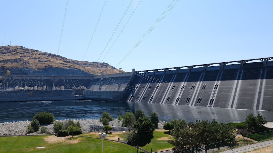 A large dam with water flowing over its spillway, a river below, and a hilly landscape in the background.